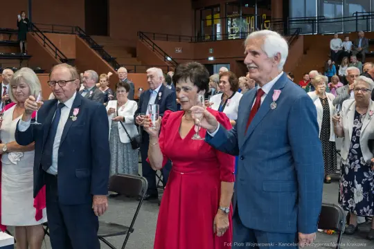 Uczestnicy wydarzenia wznoszą toast, ubrani w eleganckie stroje, z medalami na piersiach.
