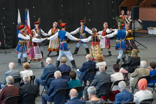 Tradycyjny taniec ludowy w kolorowych strojach, uczestnicy w ruchu, widownia w tle.