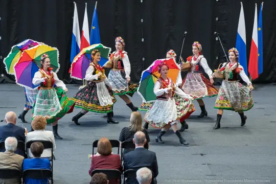 Grupa tancerzy w tradycyjnych strojach ludowych tańczy z parasolkami.