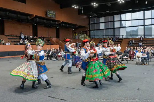 Grupa tancerzy w tradycyjnych strojach ludowych tańczy w sali, widownia obserwuje występ.