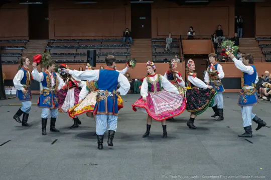 Grupa tancerzy w tradycyjnych strojach ludowych tańczy w kole, trzymając kwiaty.