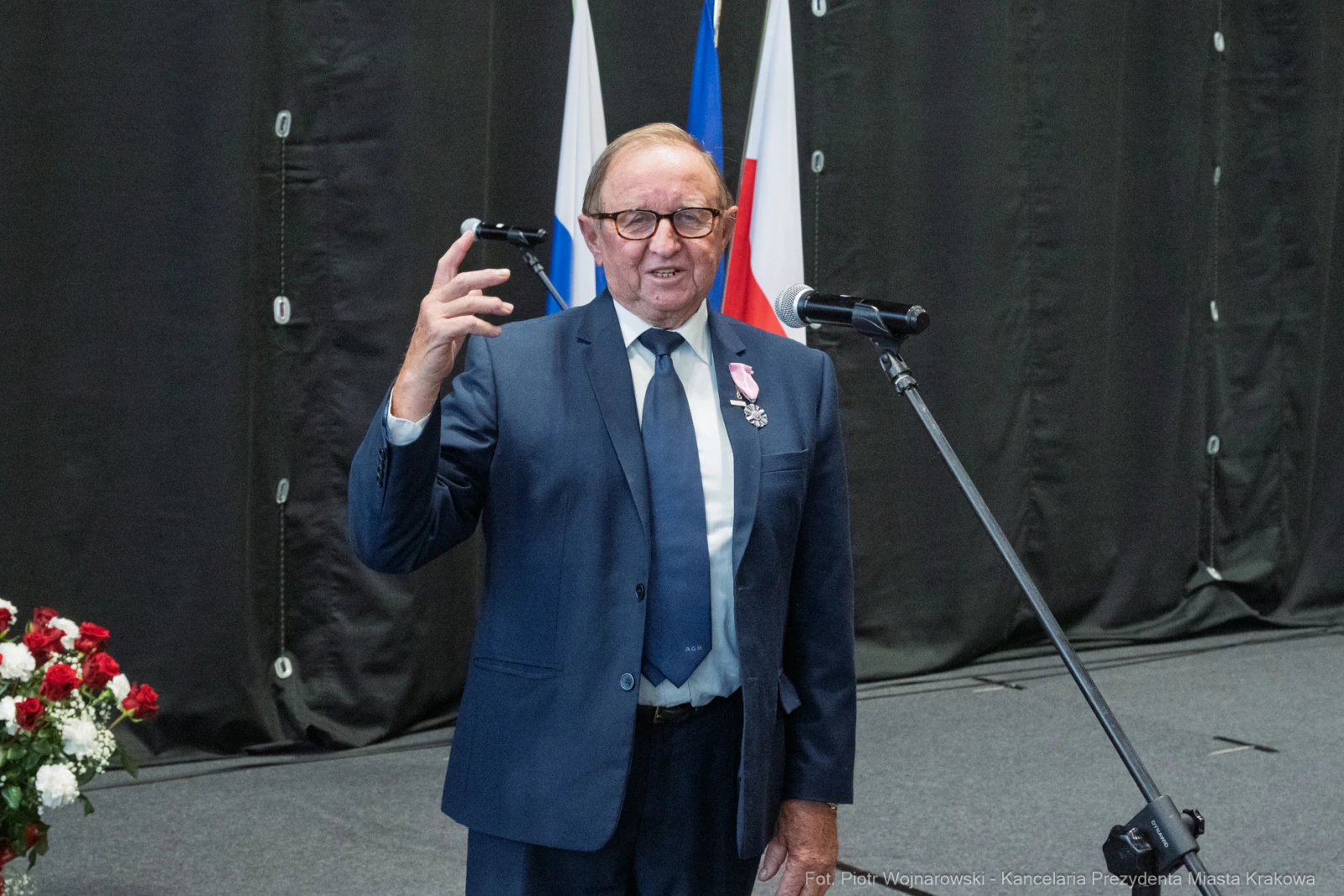 Mężczyzna w garniturze z odznaczeniem, przemawia do mikrofonu na tle flag.  Autor: Piotr Wojnarowski