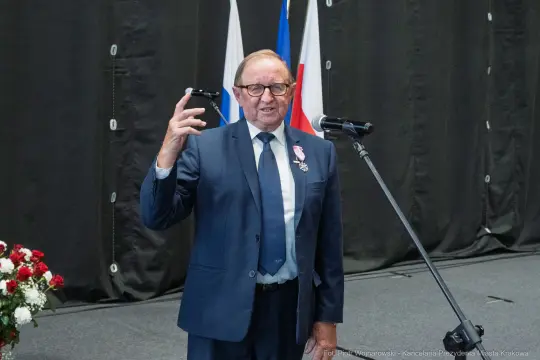 Mężczyzna w garniturze z odznaczeniem, przemawia do mikrofonu na tle flag.