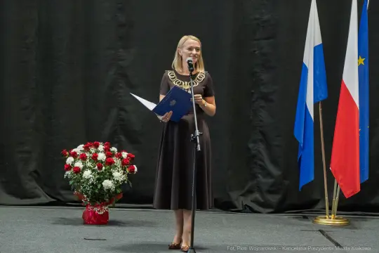Kobieta w czarnej sukience przemawia do publiczności, obok flagi i bukietu kwiatów.