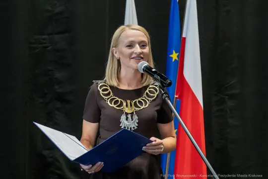 Kobieta w eleganckiej sukience z łańcuchem na szyi przemawia do mikrofonu.