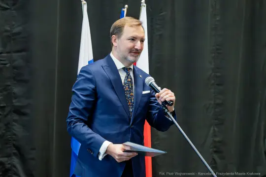 Mężczyzna w garniturze przemawia do zgromadzenia, trzymając dokument. W tle flagi.