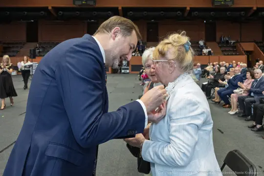 Wręczenie odznaczenia przez mężczyznę starszej kobiecie podczas uroczystości.