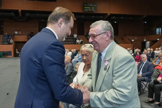 Uścisk dłoni między mężczyznami, w tle siedzący starsi ludzie.