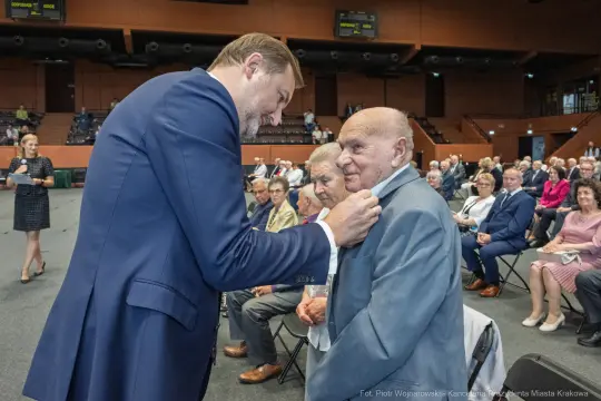Uroczystość wręczenia odznaczeń, mężczyzna przypina medal starszemu mężczyźnie.