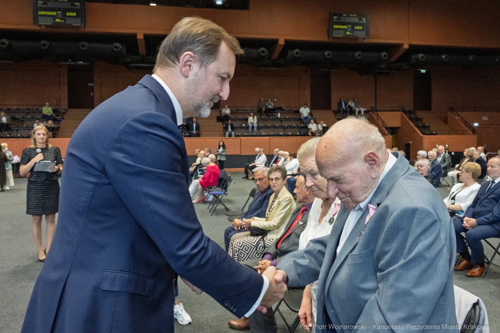 Uścisk dłoni między mężczyzną w garniturze a starszym panem w eleganckim ubraniu. W tle zgromadzenie osób.  Autor: Piotr Wojnarowski