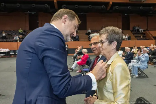 Spotkanie dwóch osób w eleganckich strojach, rozmowa w tle z innymi uczestnikami.