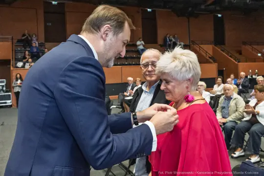 Wręczenie odznaczenia kobiecie przez mężczyznę na uroczystości w sali.