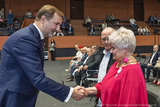 Mężczyzna w garniturze podaje rękę starszej kobiecie, w tle siedzą inni uczestnicy wydarzenia.