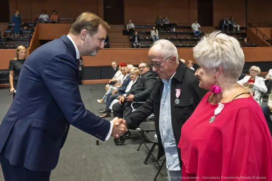 Wręczenie odznaczeń w trakcie uroczystości, mężczyzna w garniturze podaje rękę starszemu mężczyźnie.