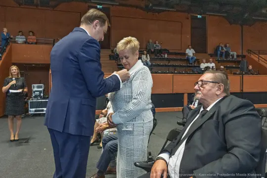 Wręczenie odznaczenia kobiecie podczas uroczystości w sali. Mężczyzna w garniturze przypina medal.
