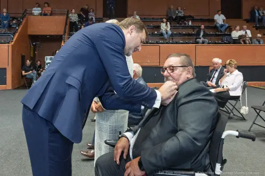 Mężczyzna w garniturze przypina odznaczenie starszemu mężczyźnie na wózku inwalidzkim.