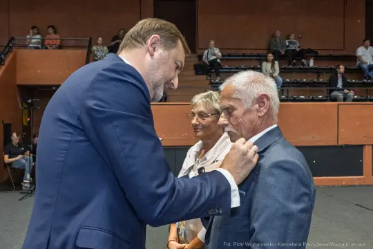 Mężczyzna przypina odznaczenie do marynarki starszego mężczyzny, obok stoi kobieta.