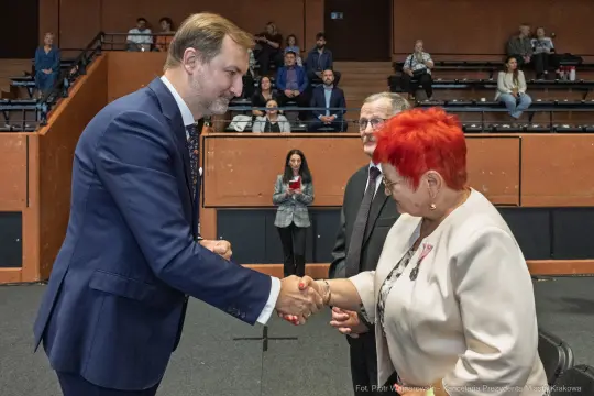 Uścisk dłoni między mężczyzną a kobietą, w tle widoczni inni uczestnicy wydarzenia.