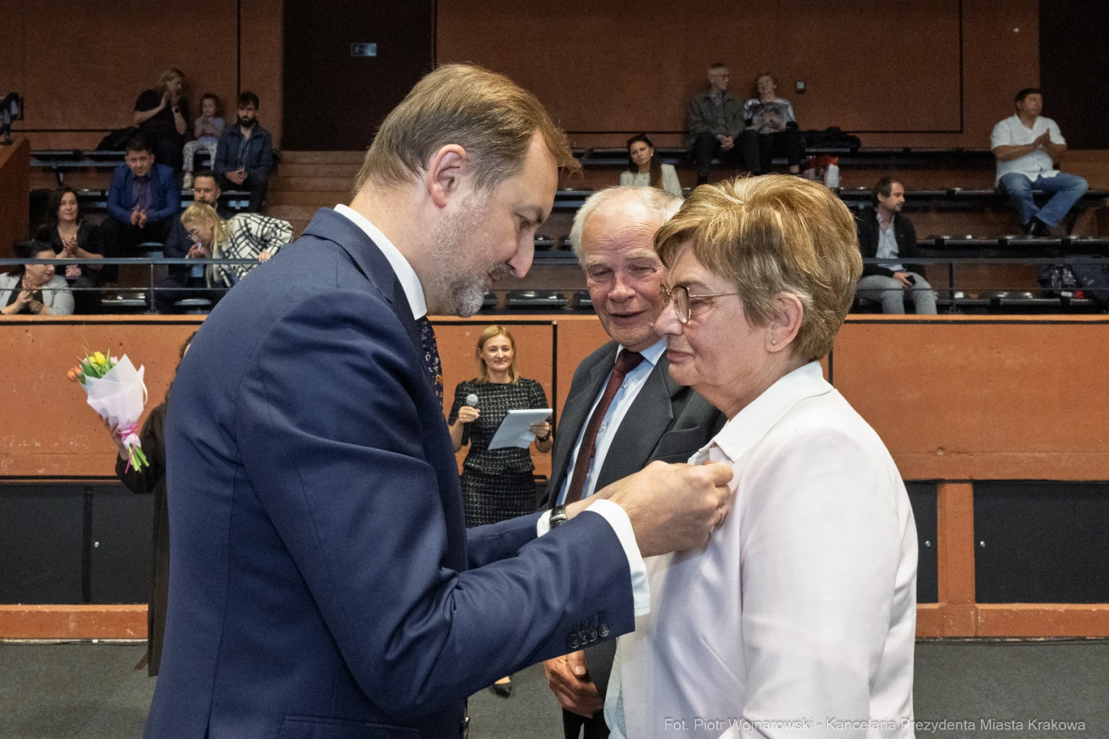 Wręczenie odznaczenia przez mężczyznę w garniturze kobiecie w białej marynarce. W tle widownia.  Autor: Piotr Wojnarowski