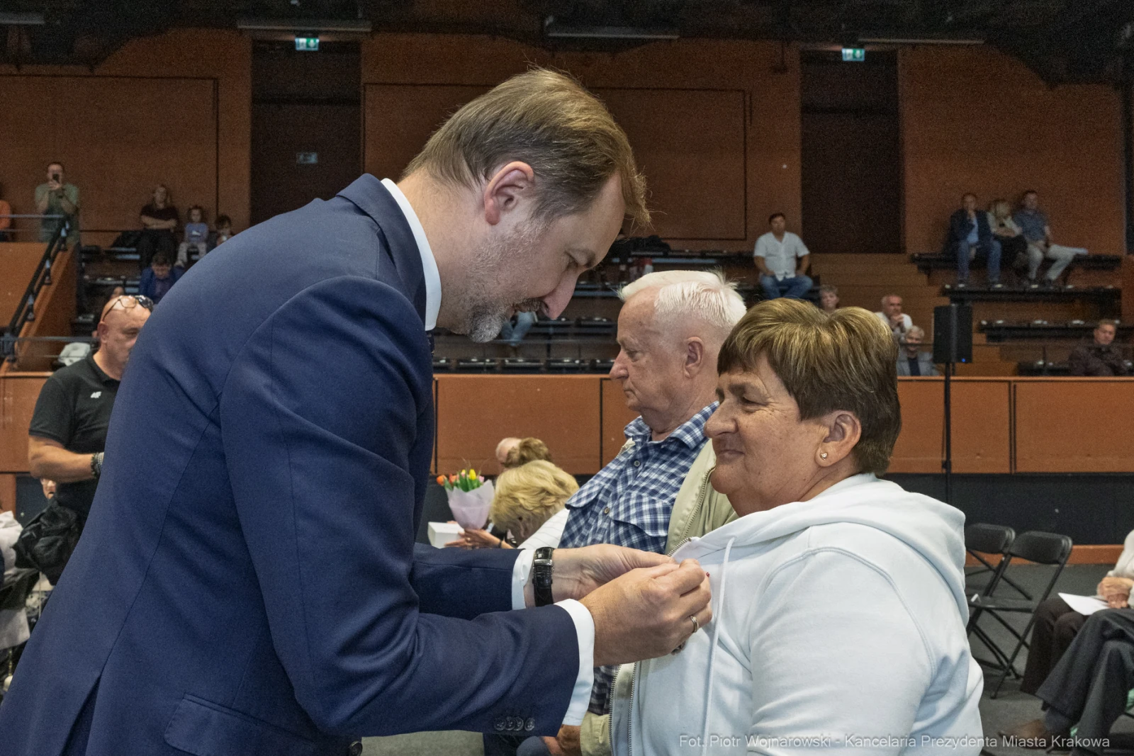 Mężczyzna przypina odznakę kobiecie podczas uroczystości w sali. W tle widoczni inni uczestnicy.  Autor: Piotr Wojnarowski