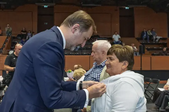 Mężczyzna przypina odznakę kobiecie podczas uroczystości w sali. W tle widoczni inni uczestnicy.