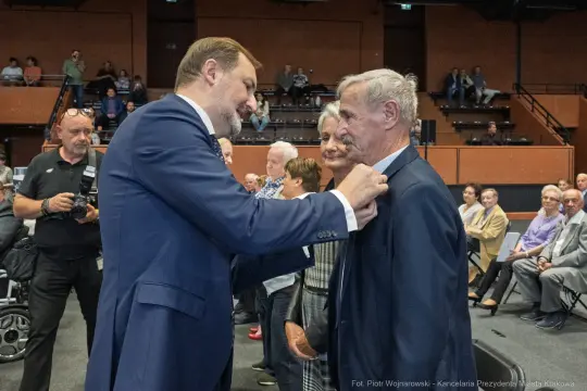 Mężczyzna przypina odznaczenie innemu mężczyźnie podczas uroczystości. W tle zgromadzeni goście.