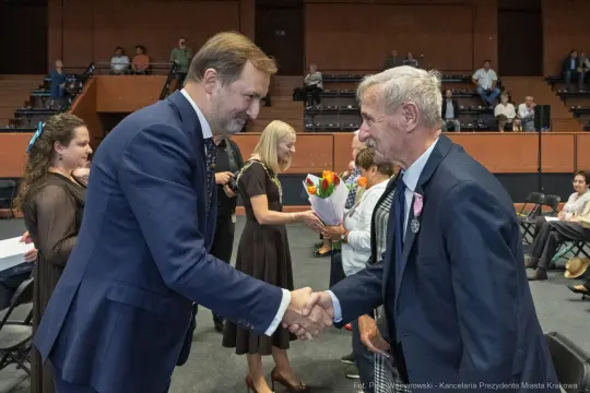 Uścisk dłoni między mężczyznami podczas uroczystości wręczenia nagród. W tle goście z kwiatami.
