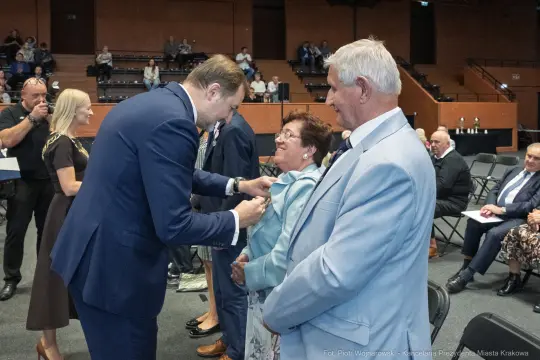 Wręczenie odznaczenia przez mężczyznę starszej kobiecie w towarzystwie mężczyzny w jasnym garniturze.