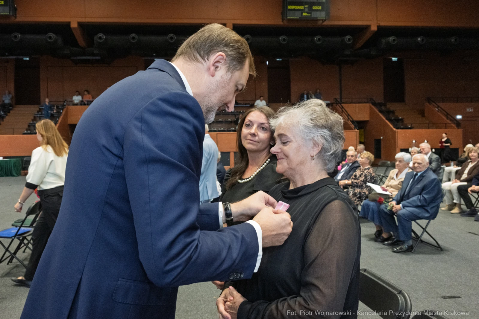 Wręczenie odznaczenia kobiecie przez mężczyznę w formalnym stroju, w tle widownia.  Autor: Piotr Wojnarowski