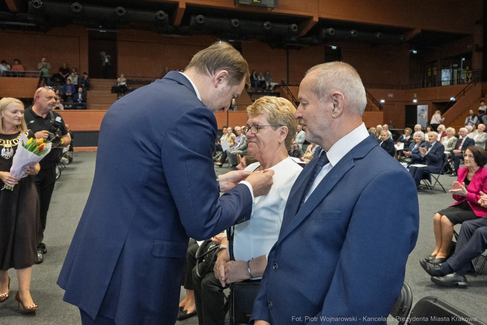 Wręczenie odznaczenia kobiecie podczas uroczystości w sali. Obecni goście w tle.  Autor: Piotr Wojnarowski