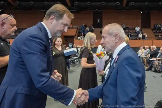 Uścisk dłoni dwóch mężczyzn podczas ceremonii wręczenia odznaczeń. W tle widoczni goście.