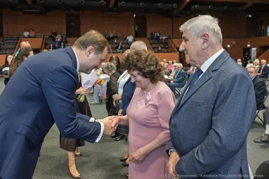 Uroczystość wręczenia nagród, osoby w formalnych strojach, uściski dłoni.