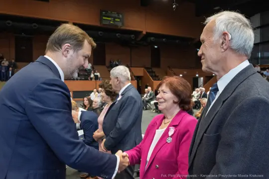 Uścisk dłoni między mężczyzną a kobietą w sali konferencyjnej, w tle zgromadzeni goście.