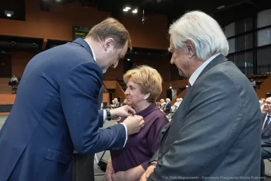 Wręczenie odznaczenia przez mężczyznę starszej kobiecie w obecności drugiego mężczyzny.