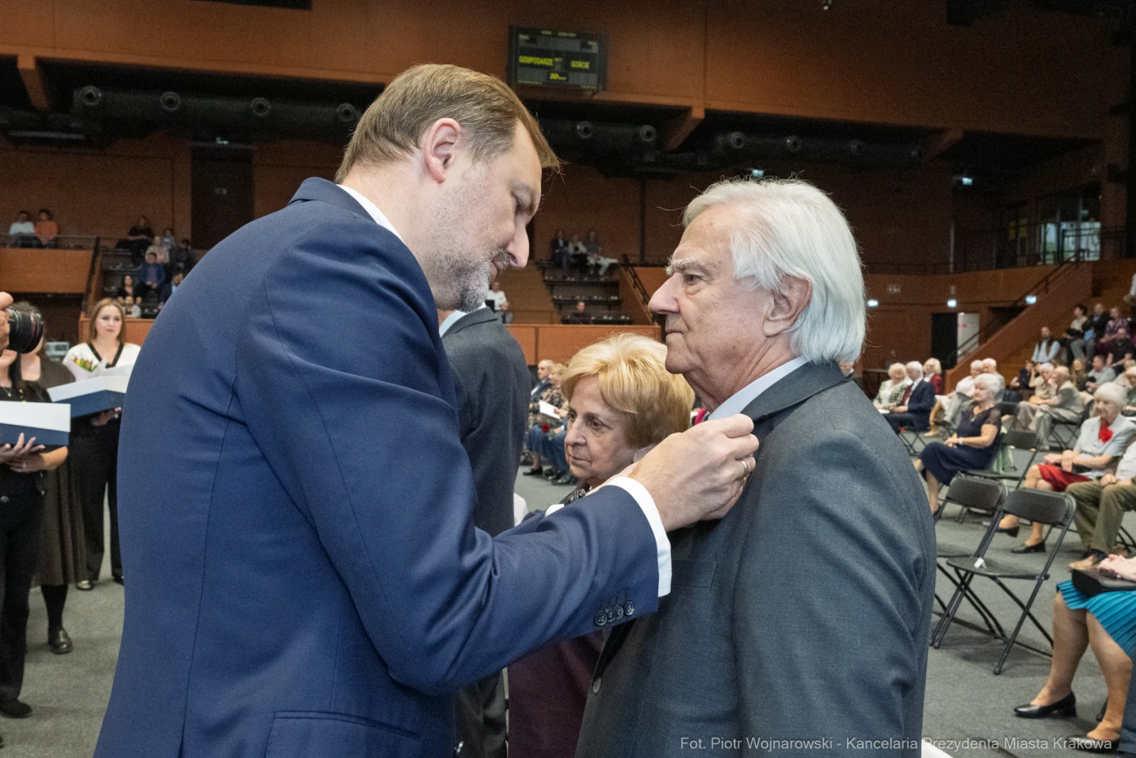 Wręczenie odznaczenia przez mężczyznę starszemu mężczyźnie w obecności kobiety. W tle zgromadzenie ludzi.  Autor: Piotr Wojnarowski