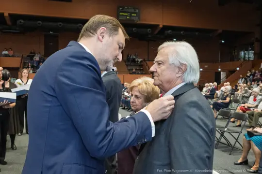 Wręczenie odznaczenia przez mężczyznę starszemu mężczyźnie w obecności kobiety. W tle zgromadzenie ludzi.