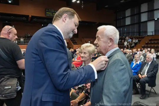 Mężczyzna przypina odznaczenie starszemu mężczyźnie w towarzystwie innych osób.