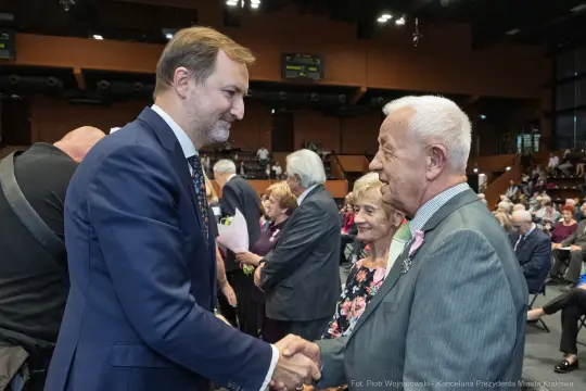 Spotkanie dwóch mężczyzn, jeden z nich uścisnął dłoń drugiego, w tle starsi ludzie.