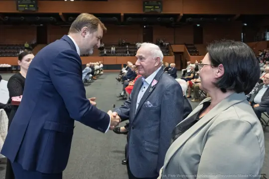 Mężczyzna w garniturze podaje rękę starszemu mężczyźnie w marynarce, obok kobieta w szarym żakiecie.