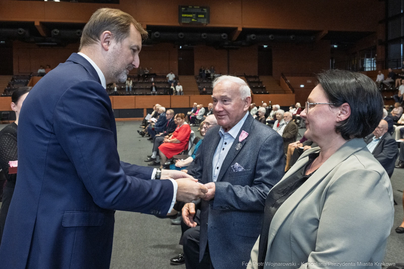 Spotkanie osób w sali, mężczyzna w garniturze wita starszego pana i kobietę.  Autor: Piotr Wojnarowski