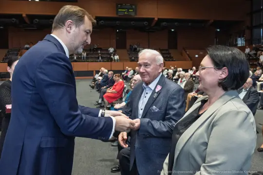 Spotkanie osób w sali, mężczyzna w garniturze wita starszego pana i kobietę.