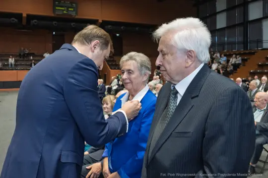 Wręczenie odznaczenia przez mężczyznę starszej parze w sali pełnej ludzi.
