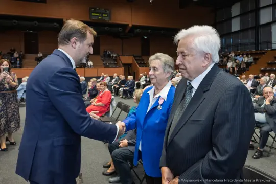 Spotkanie osób w formalnych strojach, uściski dłoni, sala wypełniona gośćmi.