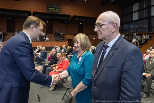 Uroczystość wręczenia odznaczeń, mężczyzna wita się z kobietą i mężczyzną w eleganckich strojach.