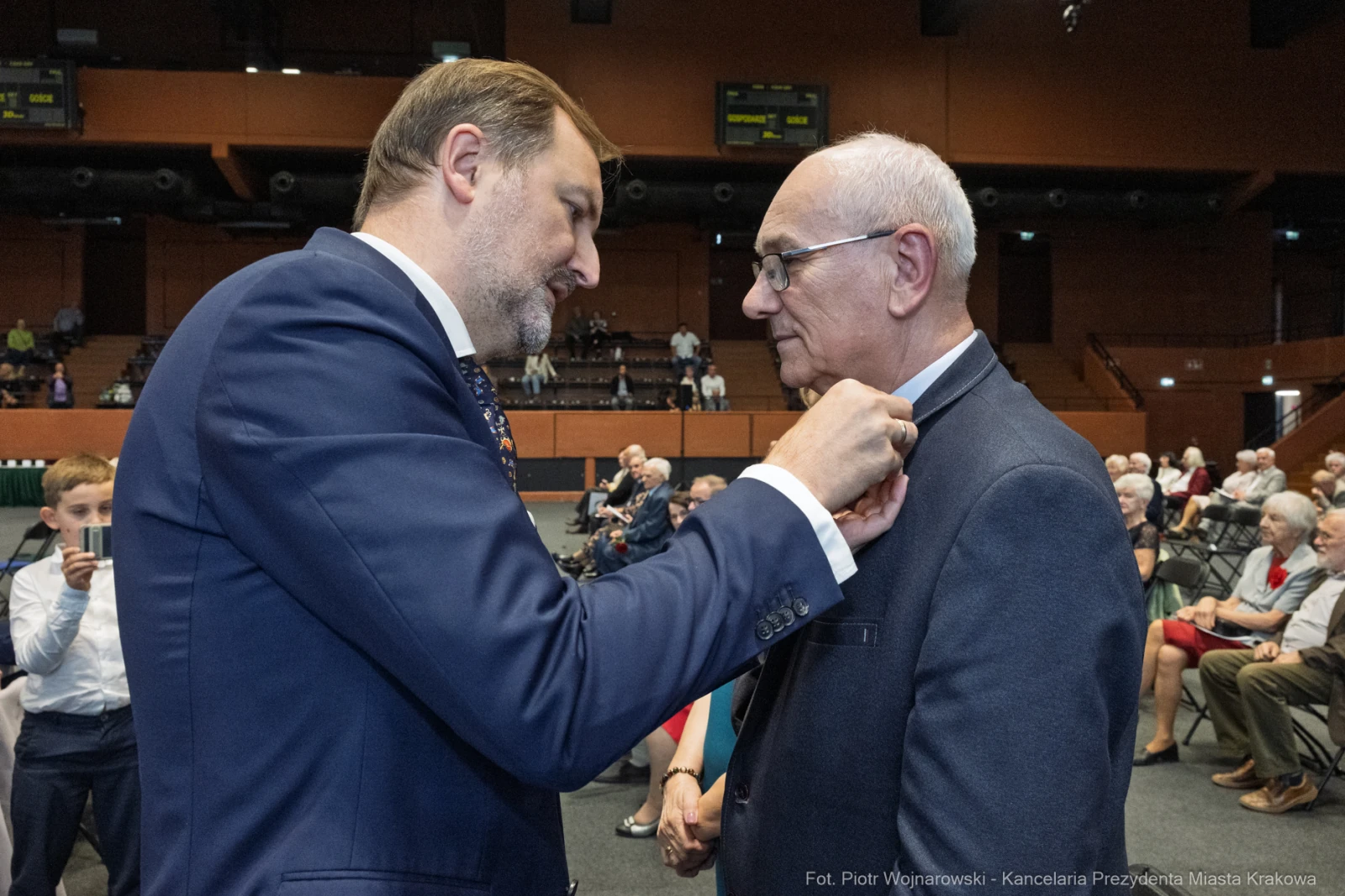 Wręczenie odznaczenia przez mężczyznę w garniturze starszemu mężczyźnie w eleganckim ubraniu.  Autor: Piotr Wojnarowski