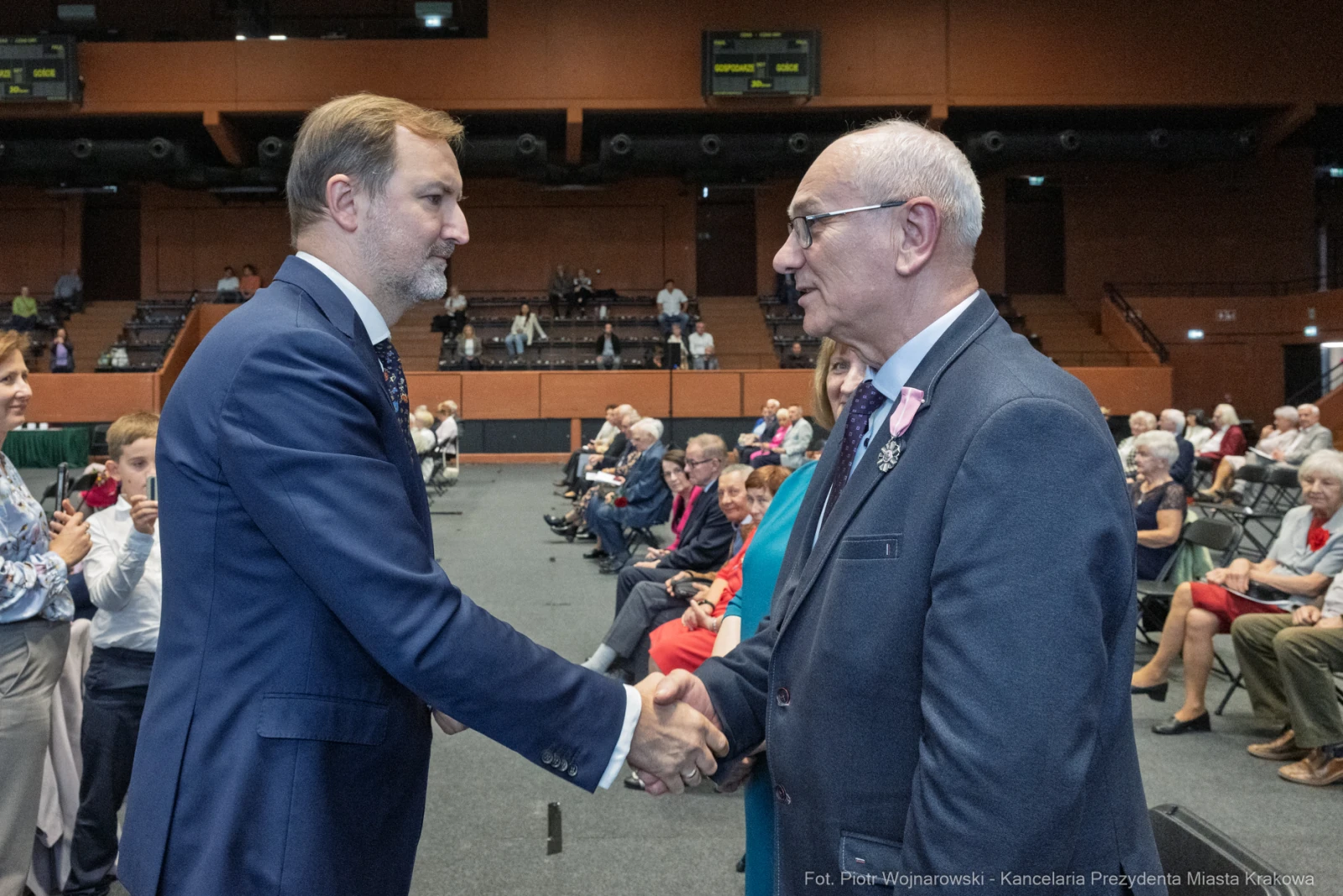 Mężczyźni uścisnęli sobie dłonie podczas wydarzenia, w tle siedzi publiczność.  Autor: Piotr Wojnarowski