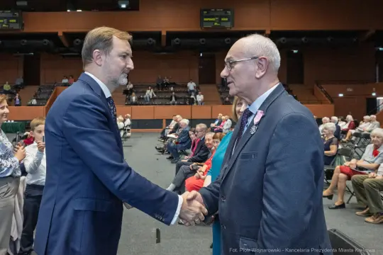 Mężczyźni uścisnęli sobie dłonie podczas wydarzenia, w tle siedzi publiczność.