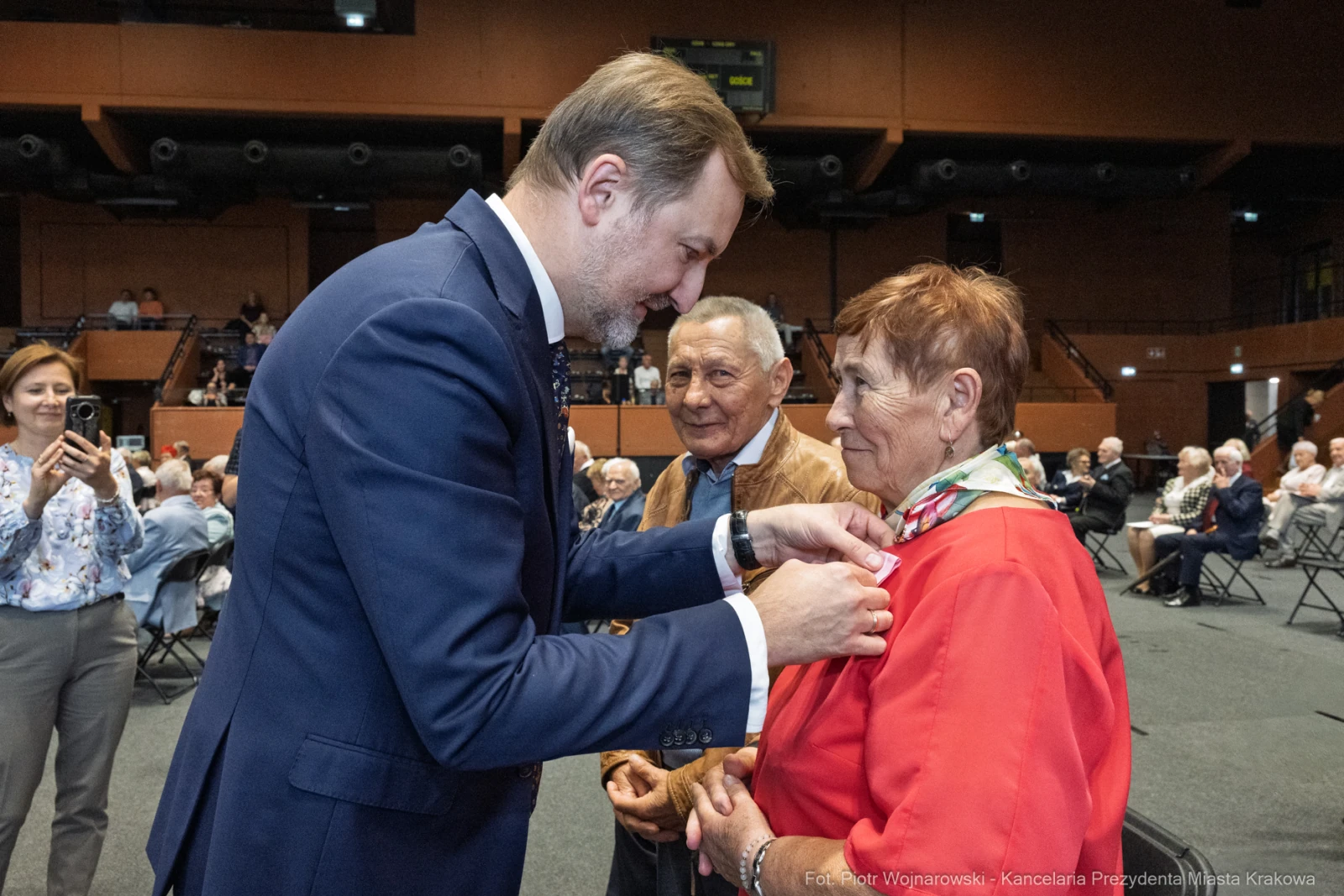 Wręczenie odznaczenia starszej kobiecie przez mężczyznę w garniturze, w tle zebrani goście.  Autor: Piotr Wojnarowski
