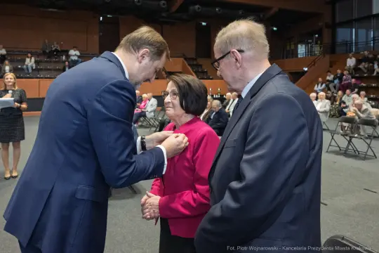 Wręczenie odznaczenia kobiecie przez mężczyznę w garniturze, obok stoi drugi mężczyzna.