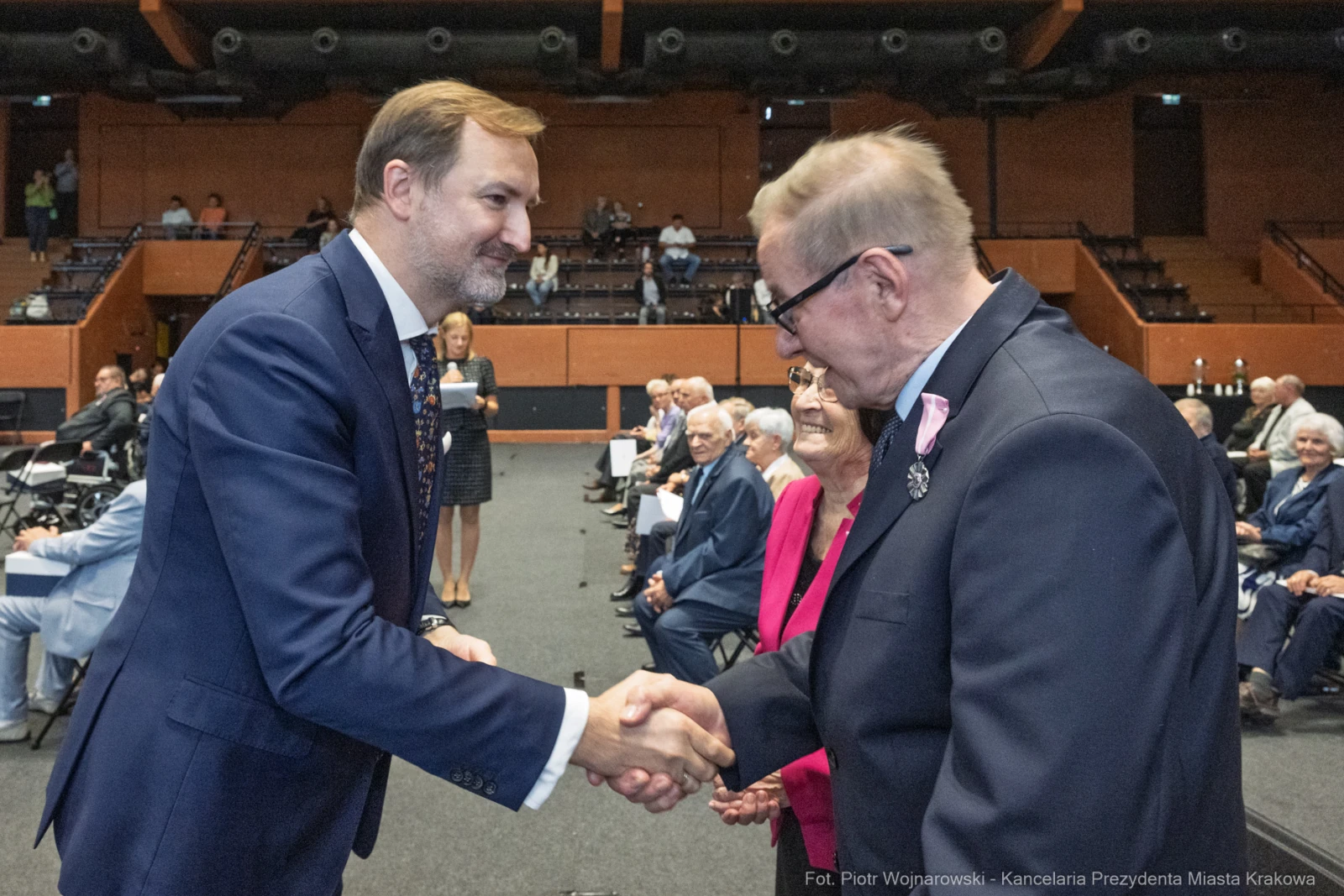 Uścisk dłoni dwóch mężczyzn w formalnym otoczeniu, w tle starsze osoby siedzące na widowni.  Autor: Piotr Wojnarowski